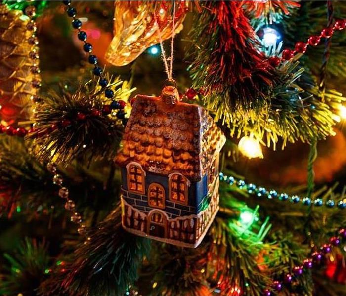 A Christmas tree ornament that looks like a house hangs on a bow amidst lights, beads, and other ornaments.
